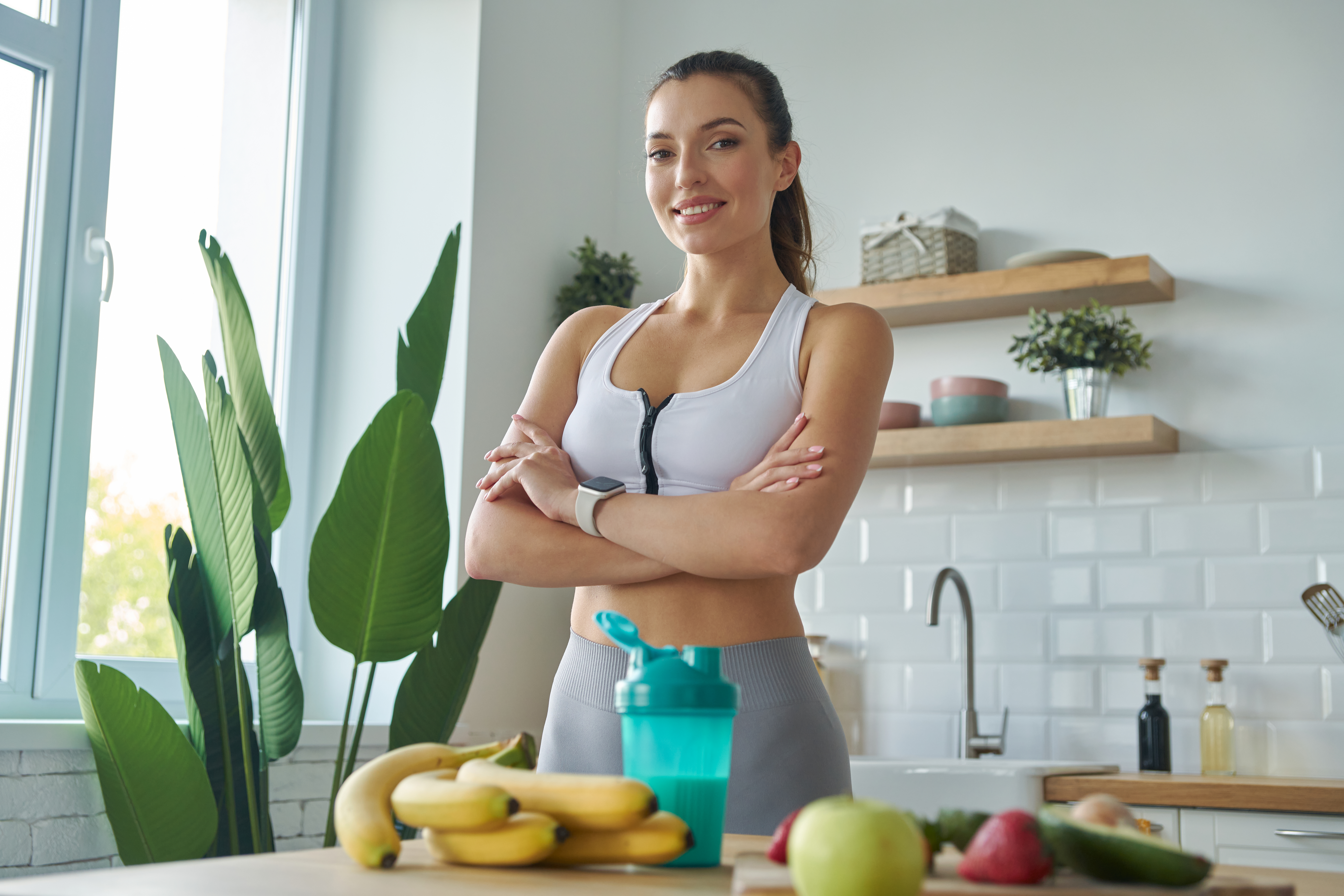 Happy young woman in sports clothing preparing protein cocktail while standing at the kitchen